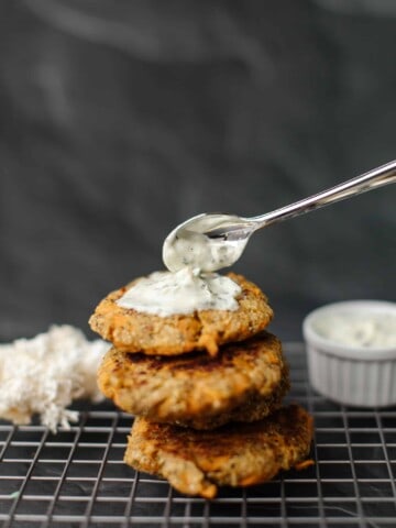 A Stack of Lion's Mane Crab Cakes on a cooling rack topped with a spoonful dollop of tartar sauce. In the background on the left is a bundle of fresh lions mane Mushroom and on the right a container of tartar sauce.