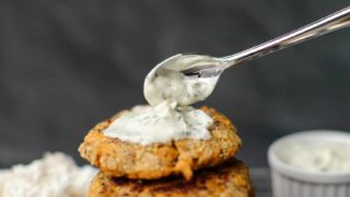 A Stack of Lion's Mane Crab Cakes on a cooling rack topped with a spoonful dollop of tartar sauce. In the background on the left is a bundle of fresh lions mane Mushroom and on the right a container of tartar sauce.