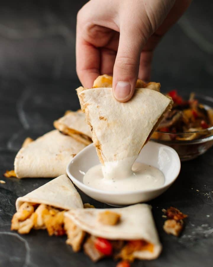 Several pieces of Mini Chicken Cheese Quesadillas surrounding a bowl of ranch dressing. A hand dipping a quesadilla triangle along with a glass bowl of filling.