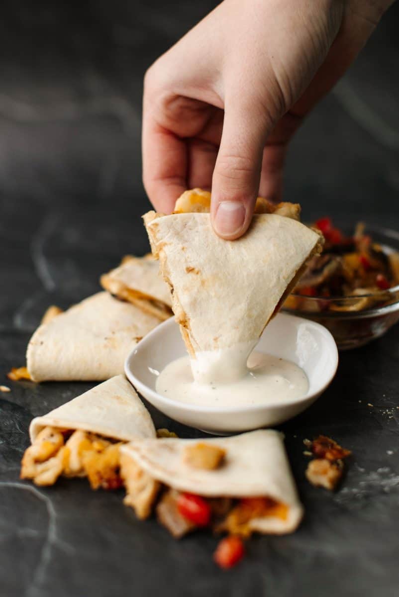 Several pieces of Mini Chicken Cheese Quesadillas surrounding a bowl of ranch dressing.  A hand dipping a quesadilla triangle along with a glass bowl of filling.