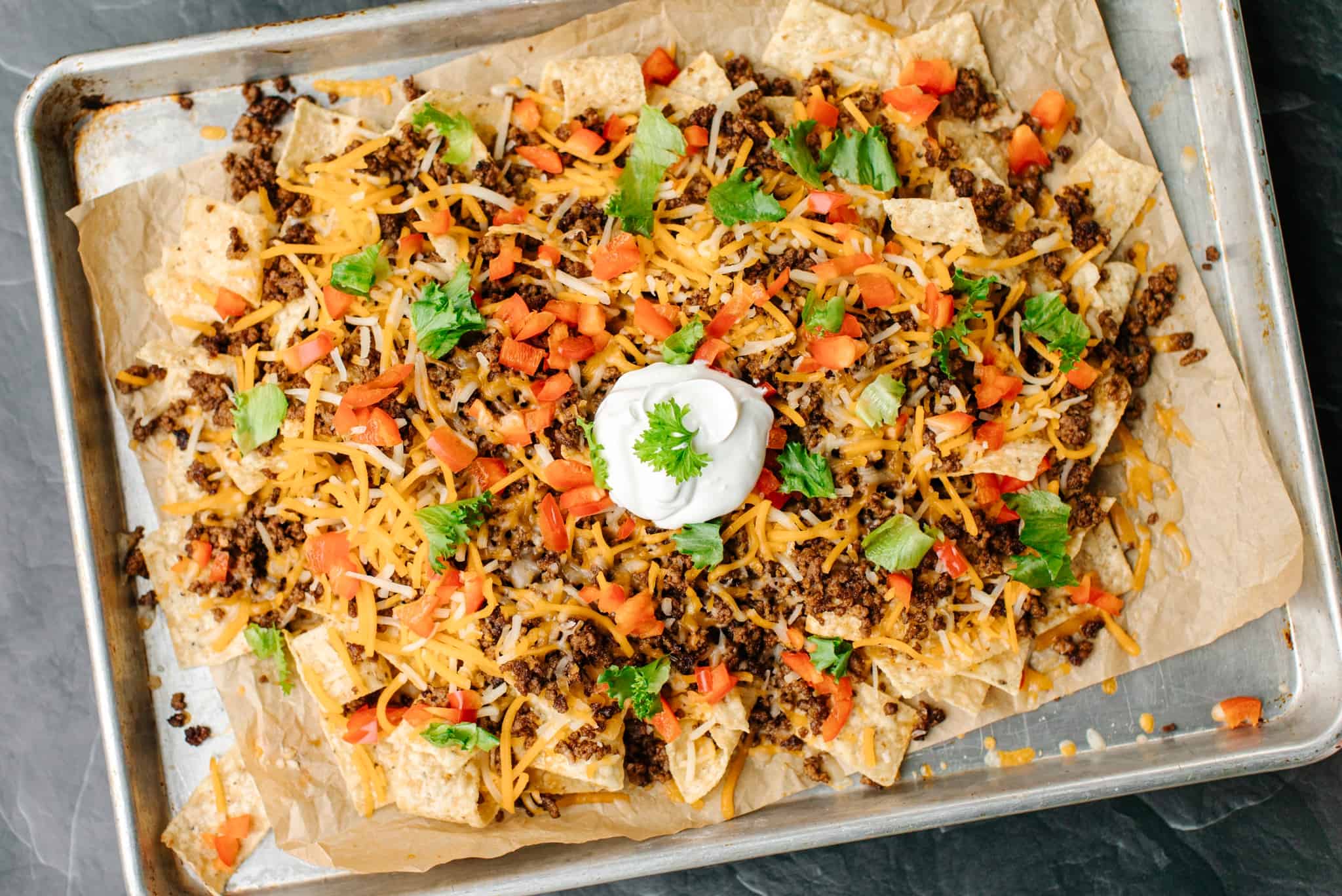 Blackstone Nachos - a sheet pan with tortilla strips, a layer of taco meat, topped with a sprinkling of shredded Colby jack cheese, diced tomatoes, and shredded lettuce.  With a dollop of sour cream right on the top middle.