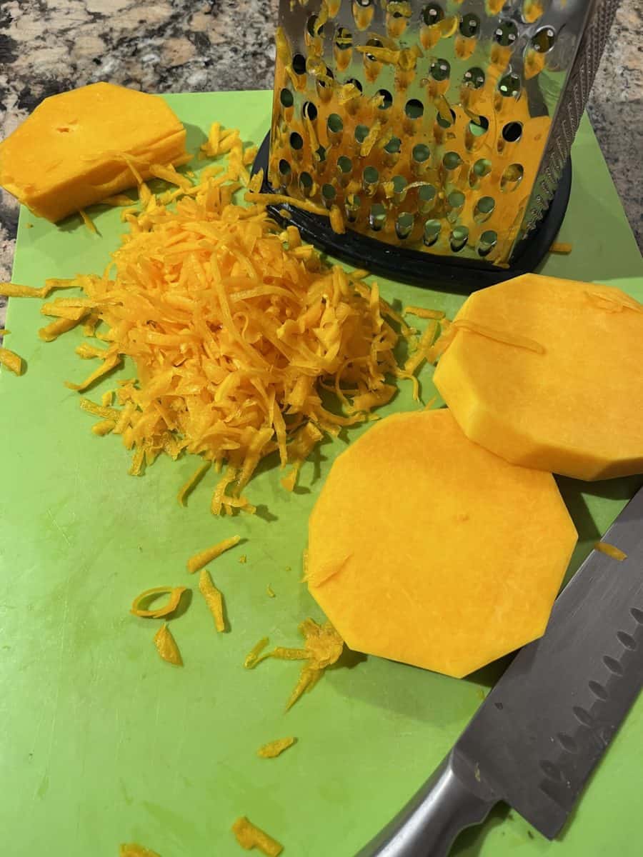 Shredded Butternut Squash on a green cutting mat with a box grater and butternut squash flat pieces.