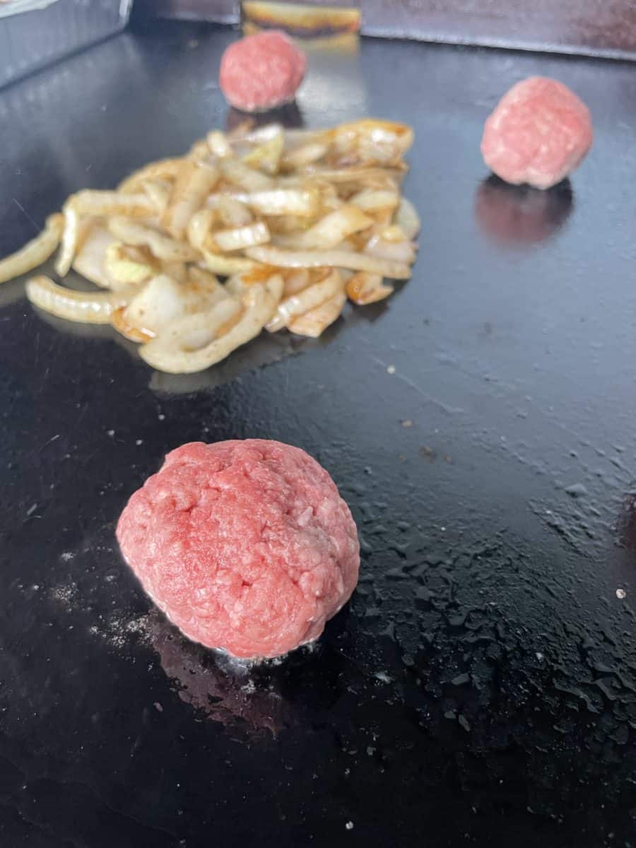 A pile of cooked onions on a flat top griddle along with a couple of round smash burger balls.