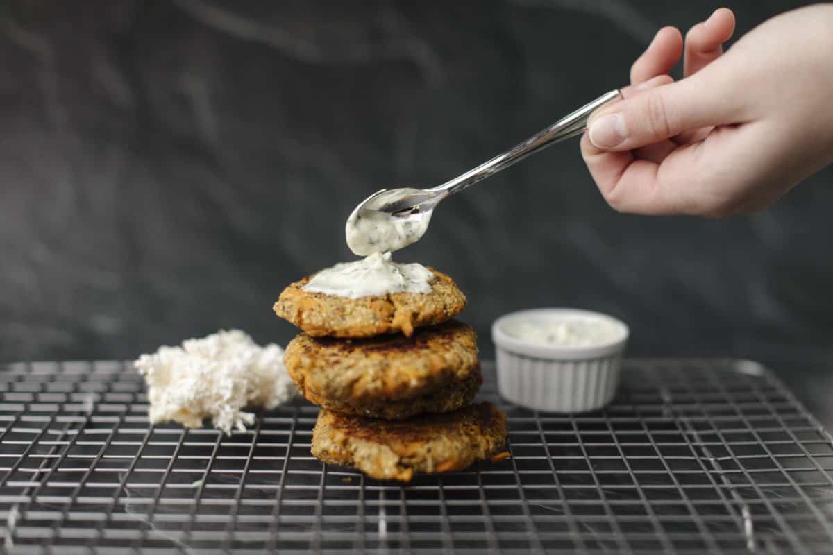 A Stack of Lion's Mane Mushroom Crab Cakes on a cooling rack topped with a spoonful dollop of tartar sauce. In the background on the left is a bundle of fresh lions mane Mushroom and on the right a container of tartar sauce.