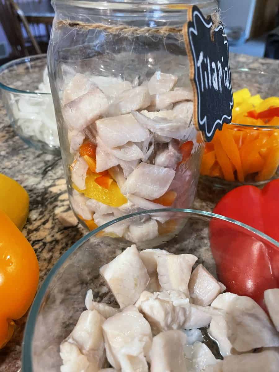 Layering the pickled tilapia in a quart jar surrounded by the whole and sliced onions and peppers and a bowl of brined tilapia pieces.