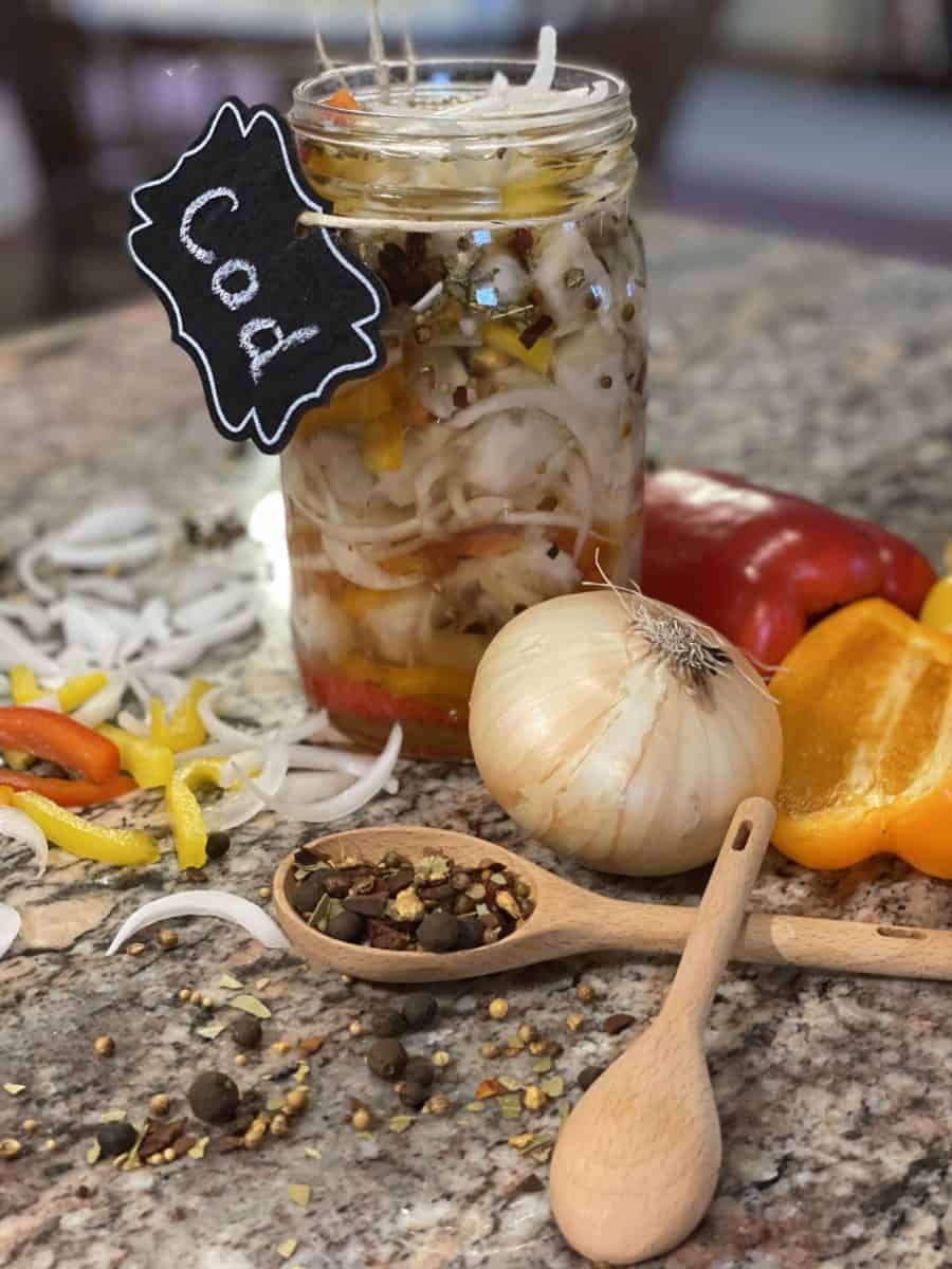 Pickled Fish with Cod - Quart glass jar full of pickled fish surrounded by sliced onion, peppers, a small wooden spoon full of pickling spice and a large onion, with half peppers.