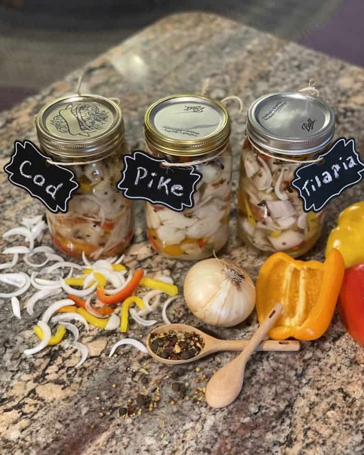 Pickled Cod, Pickled Pike and Pickled Tilapia -Quart glasses jar full of pickled fish surrounded by sliced onion, peppers, a small wooden spoon full of pickling spice and a large onion, with half peppers.