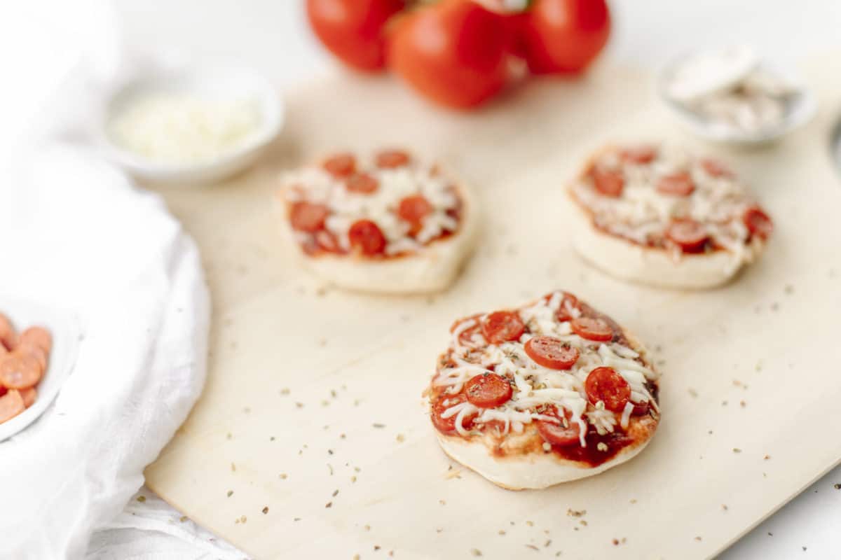 Mini Pizzas that have been cooked on a Blackstone Griddle and displayed on a wooden pizza peel with a small bowl of shredded cheese and a cluster of tomatoes.