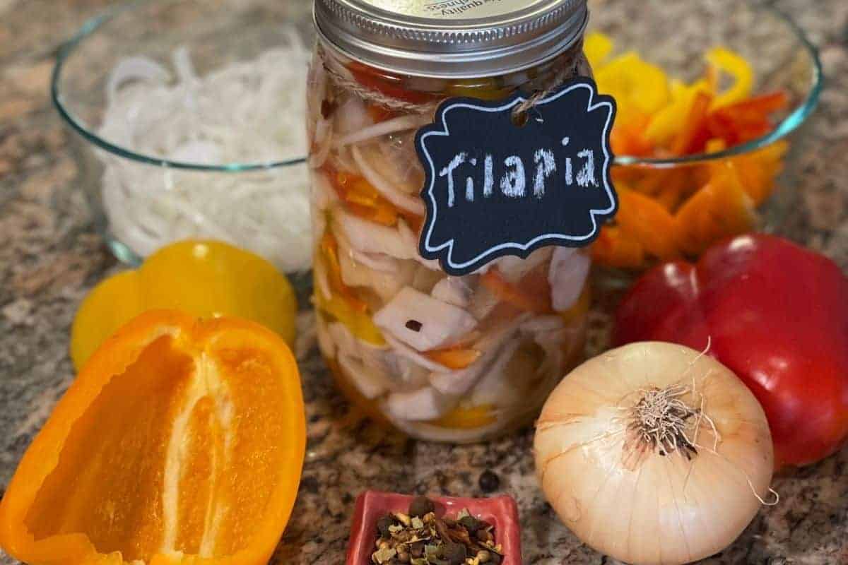 Pickled Tilapia in a quart glass jar surrounded by the whole and sliced onions and peppers with a small container of pickling spice.