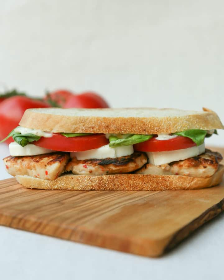 Chicken Caprese Sandwich on a wooden cutting board with a bunch of whole tomatoes in the background.