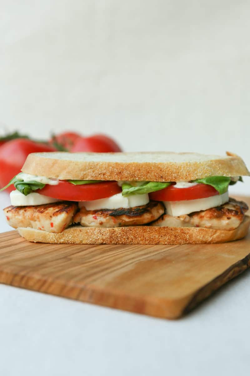 Chicken Caprese Sandwich on a wooden cutting board with a bunch of whole tomatoes in the background.