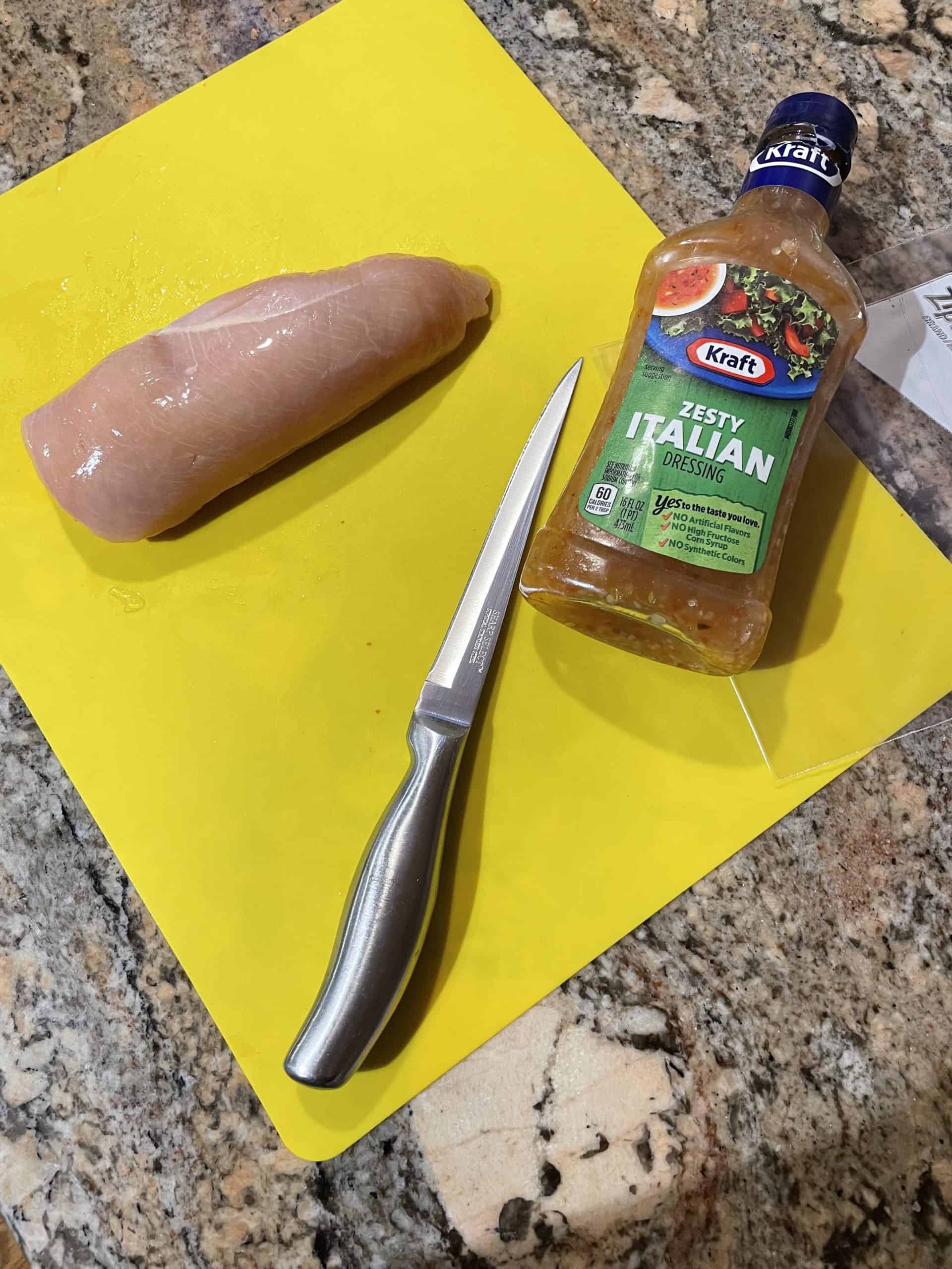Chicken Breast, a sharp knife and Italian dressing on a yellow cutting mat.