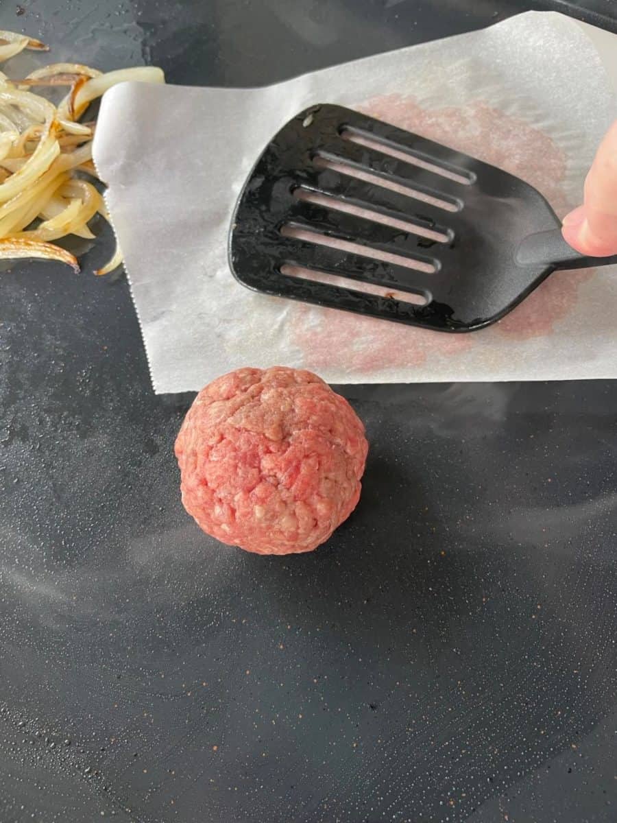 Smashing a Burger Ball on a Blackstone Griddle with a spatula along with another ball and grilled onions.