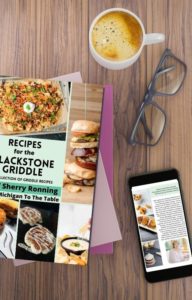 Recipes for the Blackstone Griddle Cookbook on a wooden table with a phone, glasses and a cup of coffee.