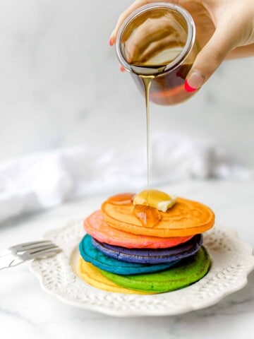 Rainbow Pancakes on the Griddle in a stack on a plate topped with a pat of butter and syrup being poured on top.