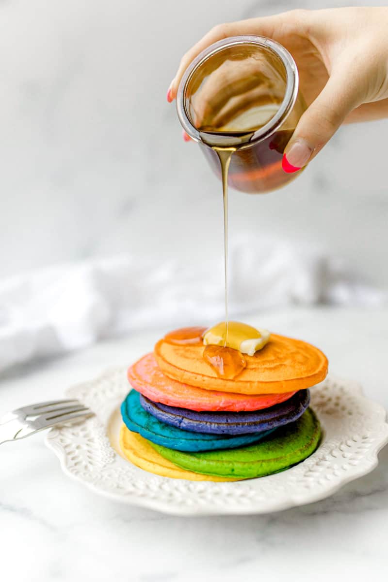 Rainbow Pancakes on the Griddle in a stack on a plate topped with a pat of butter and syrup being poured on top.