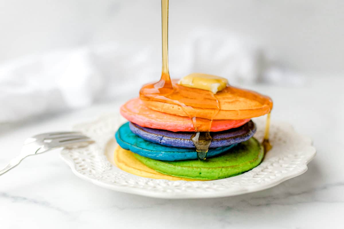 Rainbow Pancakes stacked on a plate topped with a pat of butter and syrup being poured on top.