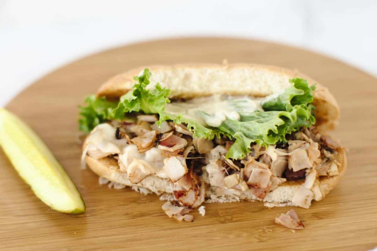 Ham, Mushroom, and Turkey Sub Sandwich topped with melted cheese, lettuce, and mayo on a wooden board alongside a pickle spear.  