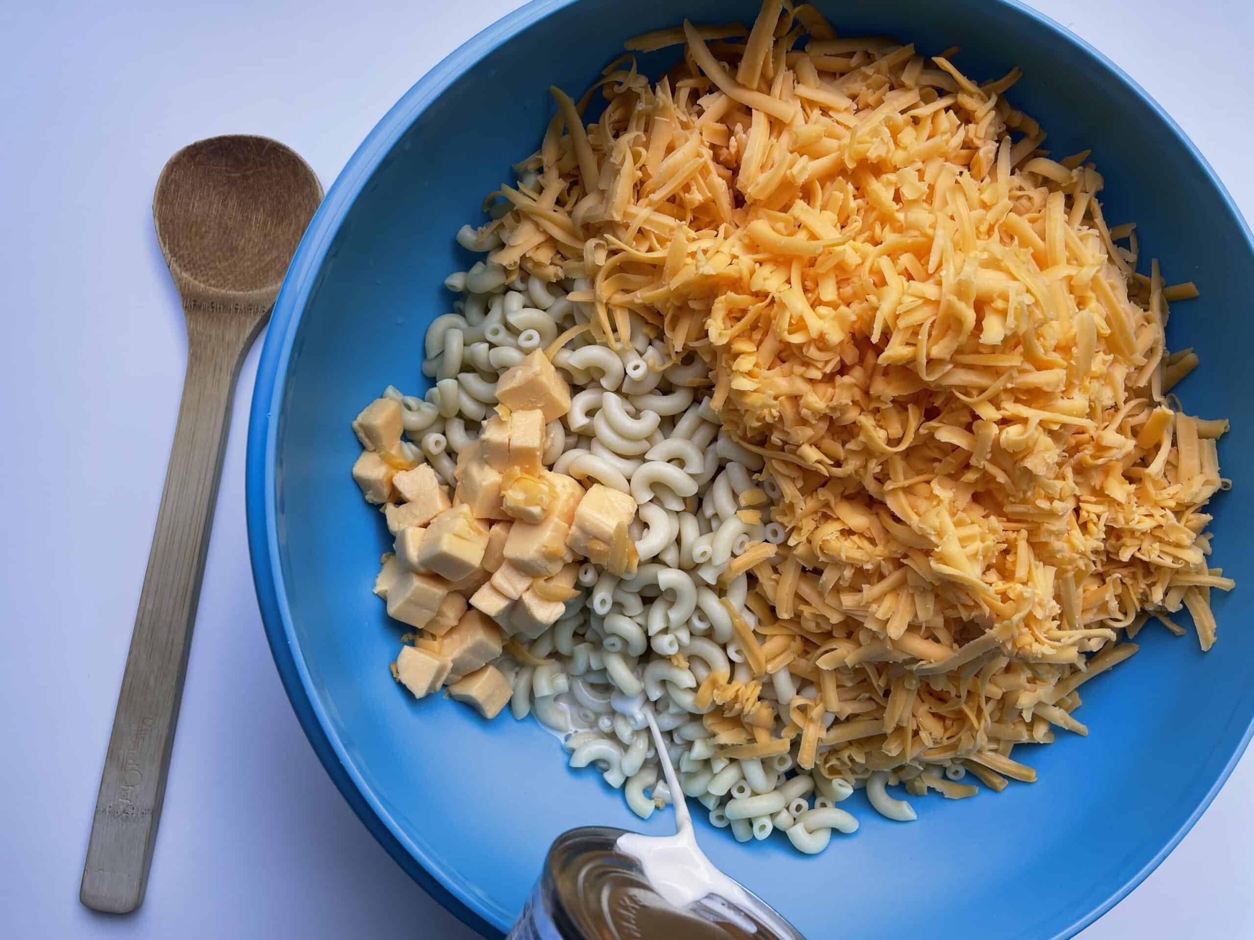 A large bowl with cooked macaroni noodles, shredded cheese and diced Velveeta cheese along with pouring canned milk. 