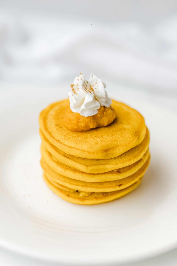 Bisquick Pumpkin Pancakes stacked up on a plate and topped with a dollop of pumpkin puree and whipped cream.