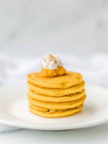 A stack of pumpkin pancakes on a plate with a dollop of whipped cream and pumpkin puree.