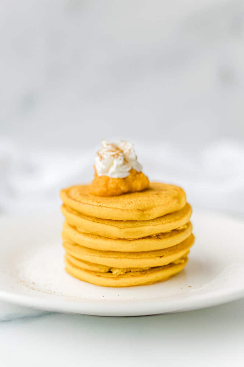 A stack of pumpkin pancakes on a plate with a dollop of whipped cream and pumpkin puree.
