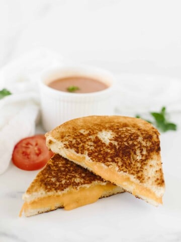 A Blackstone Grilled Cheese Sandwich cut in half and stacked along with a cup of tomato soup, slices of tomato, and a white cloth napkin.