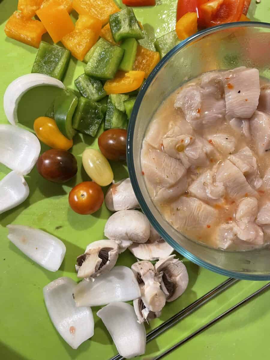Kebob Ingredients: Marinated Boneless Skinless Chicken Breast Pieces in a Bowl, Surrounded by Several Pieces of Vegetables - Mushrooms, baby tomatoes, white onion, and peppers.