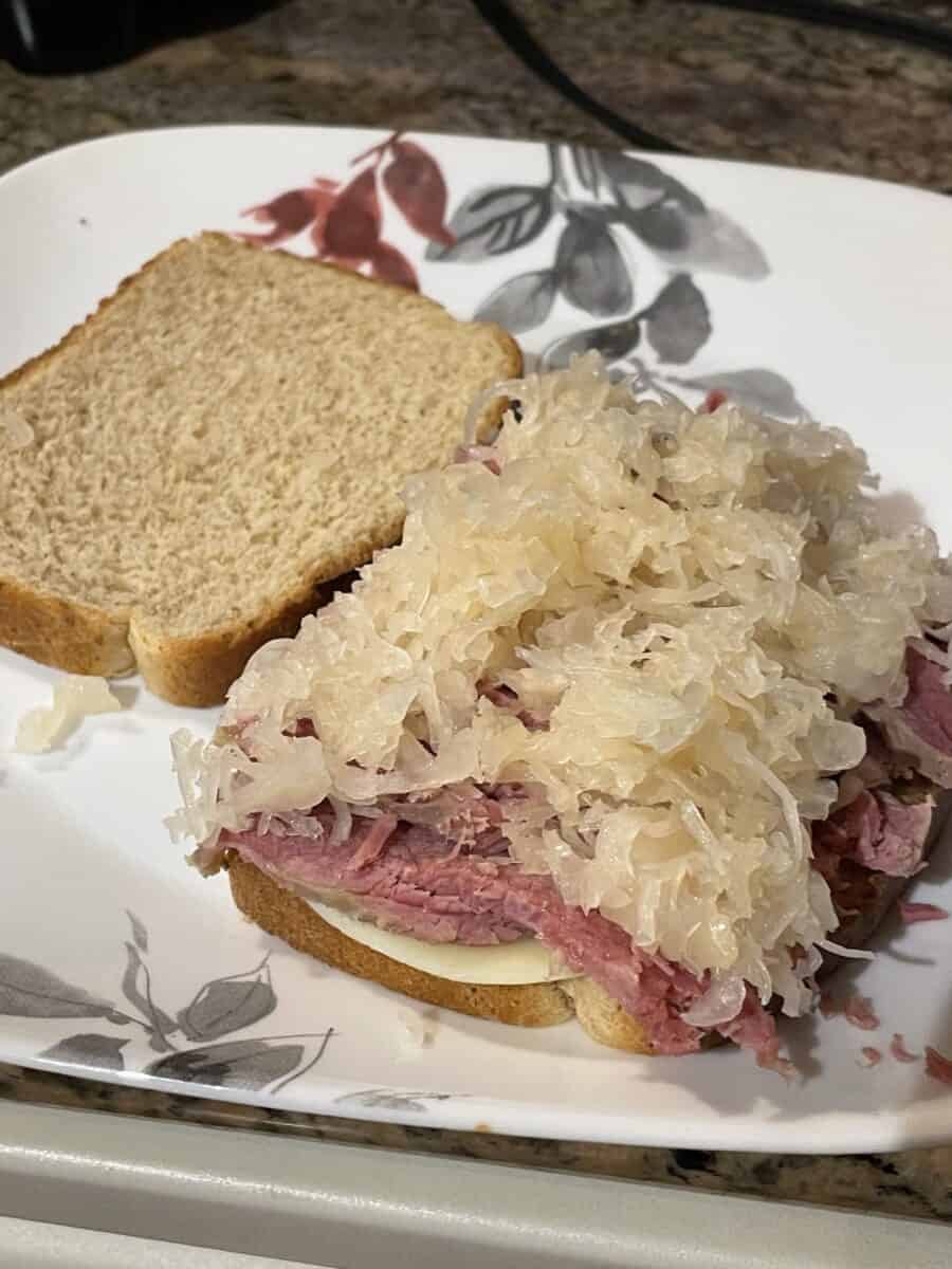 Corned Beef Sandwich made from 2 slices of bread, cheese slices, corned beef slices, and topped with sauerkraut on a plate.