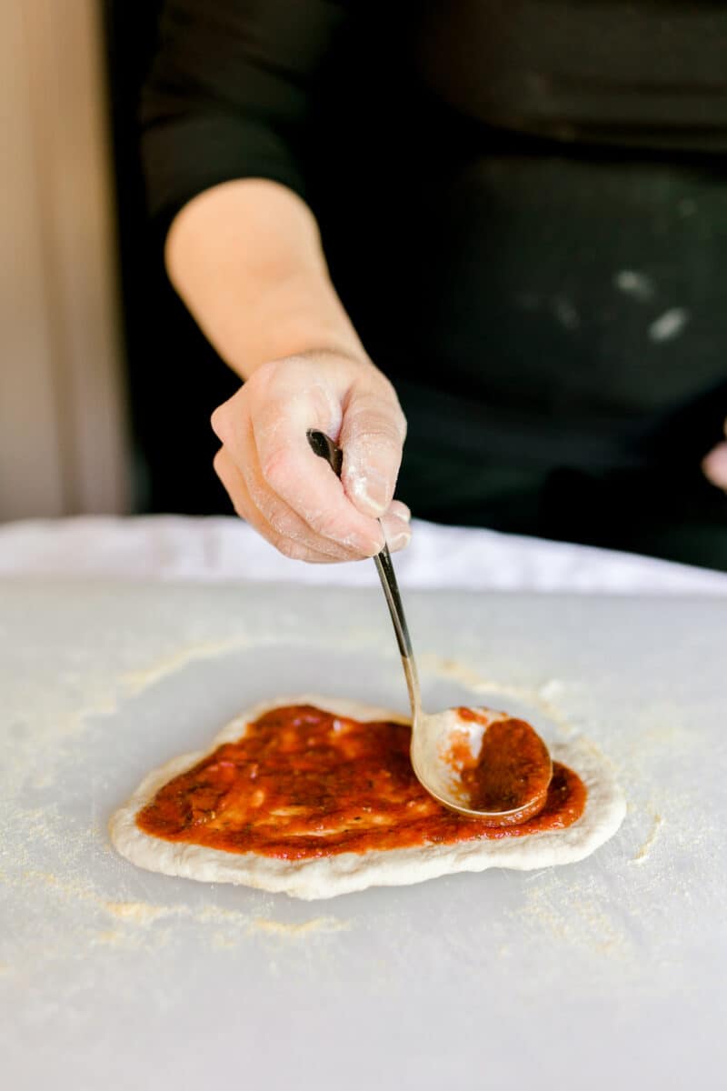 Using a Ladle to Spread Pizza Sauce onto Pizza Dough.