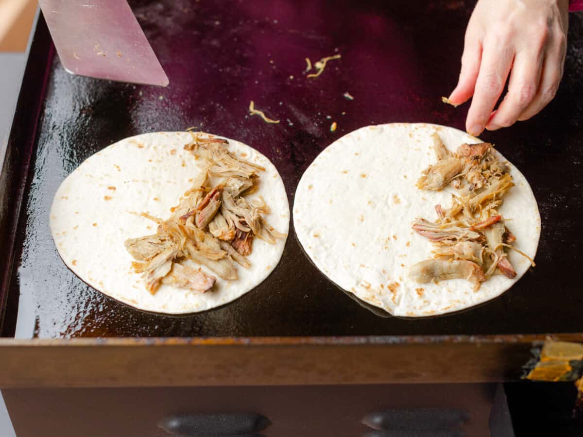 Adding the Shredded Pork on top of the tortilla shell as it is cooking on a flat top griddle.