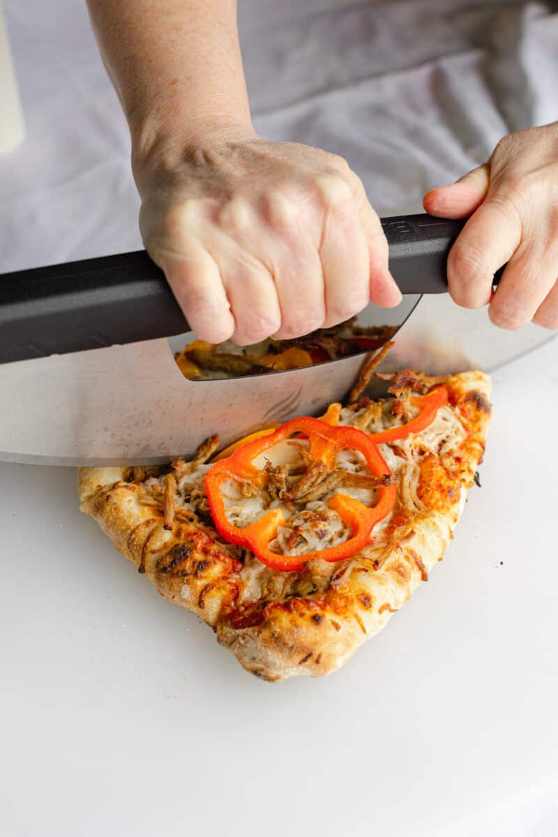 Using a Pizza Rocker to Cut a Fully Baked Shredded Pork Pizza on a Cutting Board.