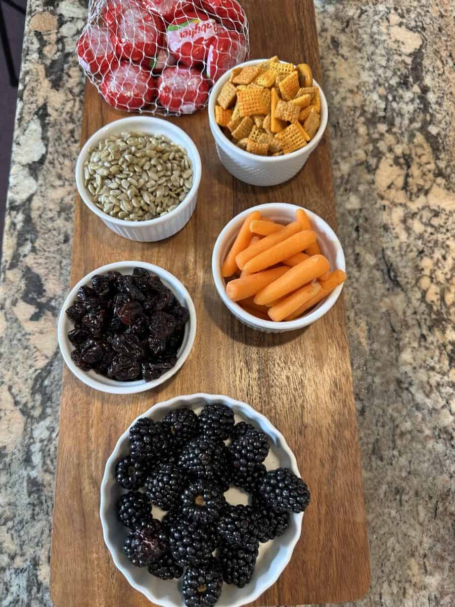 Charcuterie Board Ingredients in Small Bowls: Baby Carrots, Dried Cherries, Fresh Blackberries, Seeds, Cheddar Chex Mix, and Babybel Cheese.  