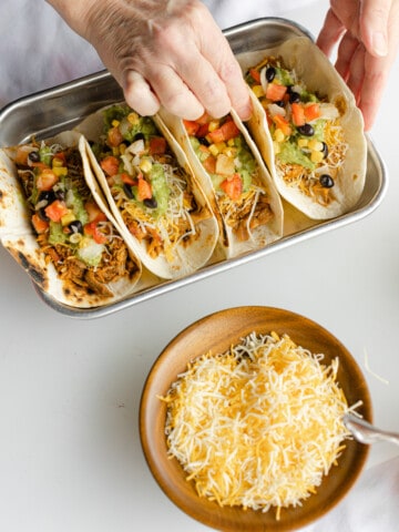4 Pulled Pork Tacos lined up on a small sheet pan with a side bowl of shredded cheese.