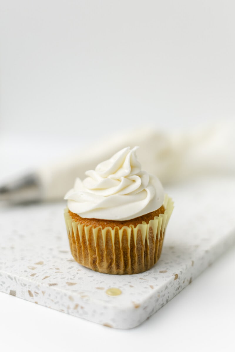 Butter Cream Cheese Frosting on top of a cupcake with a piping bag in the background.