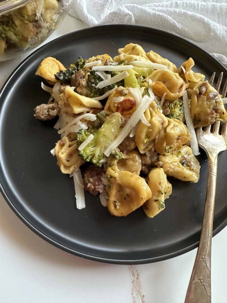 A black dinner plate with Griddle Creamy Tortellini with broccoli and sausage.