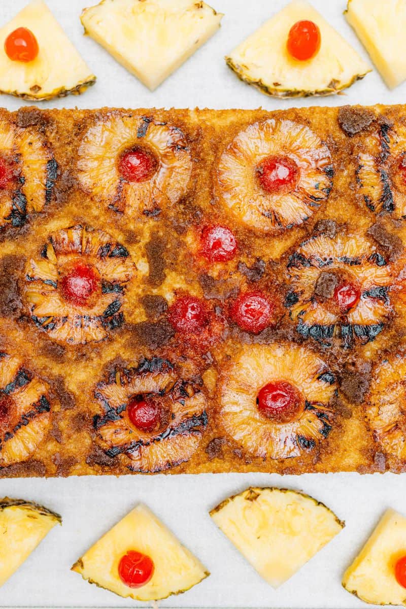 A Whole Grilled Pineapple Up-Side Down Cake surrounded by fresh wedges of pineapples and maraschino cherries